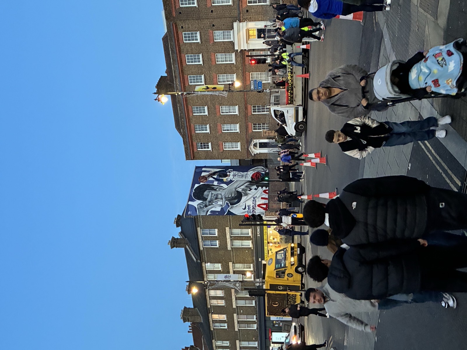 Crowds on Tottenham High Road at dusk, a footballer mural on the wall, heading to the stadium.